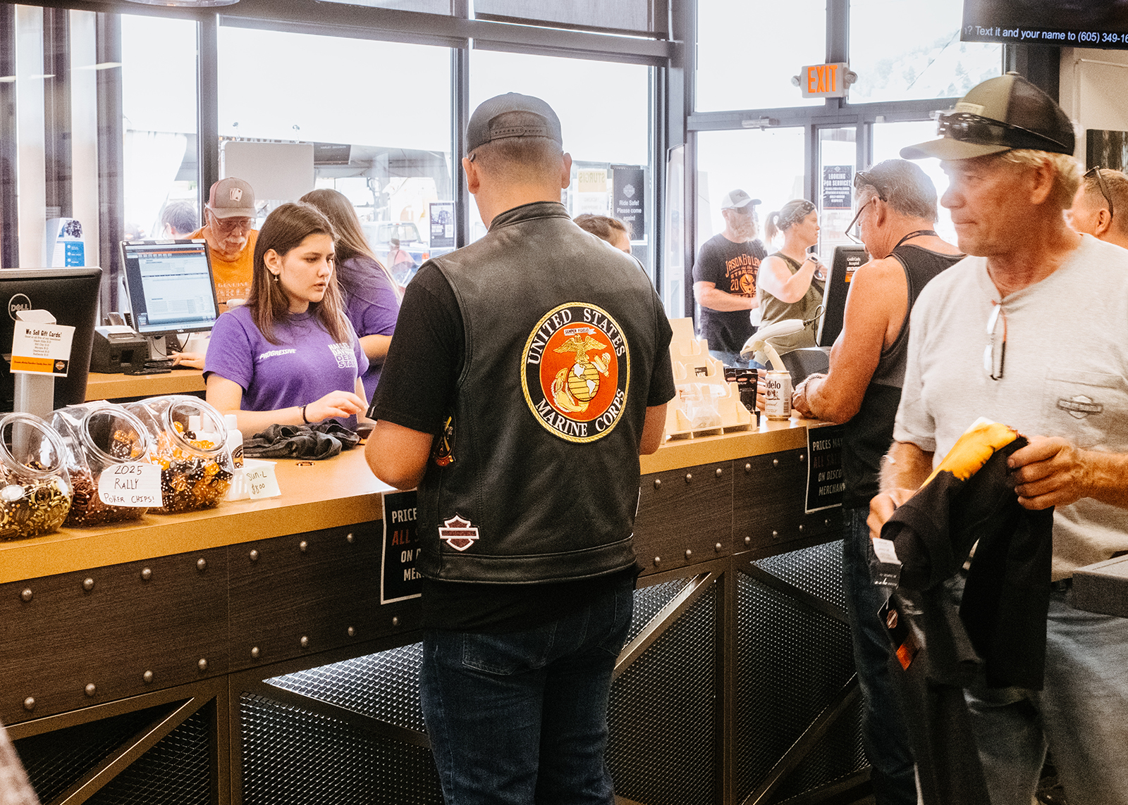 Customer wearing a US Marine Corps vest checks out at the Sturgis Harley-Davidson store counter, staff in purple shirts assist with sales.