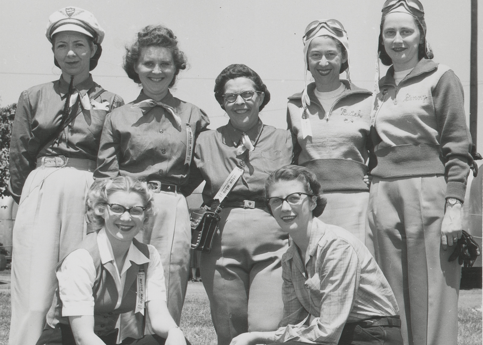 A vintage black and white photo of early Motor Maids members smiling together in uniform shirts, slacks, and caps.