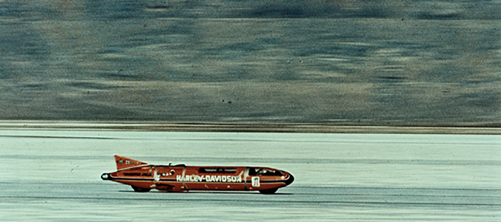 An image of the orange Sportster streamliner in action, racing across the salt flats, as the background blurs with the speed. 