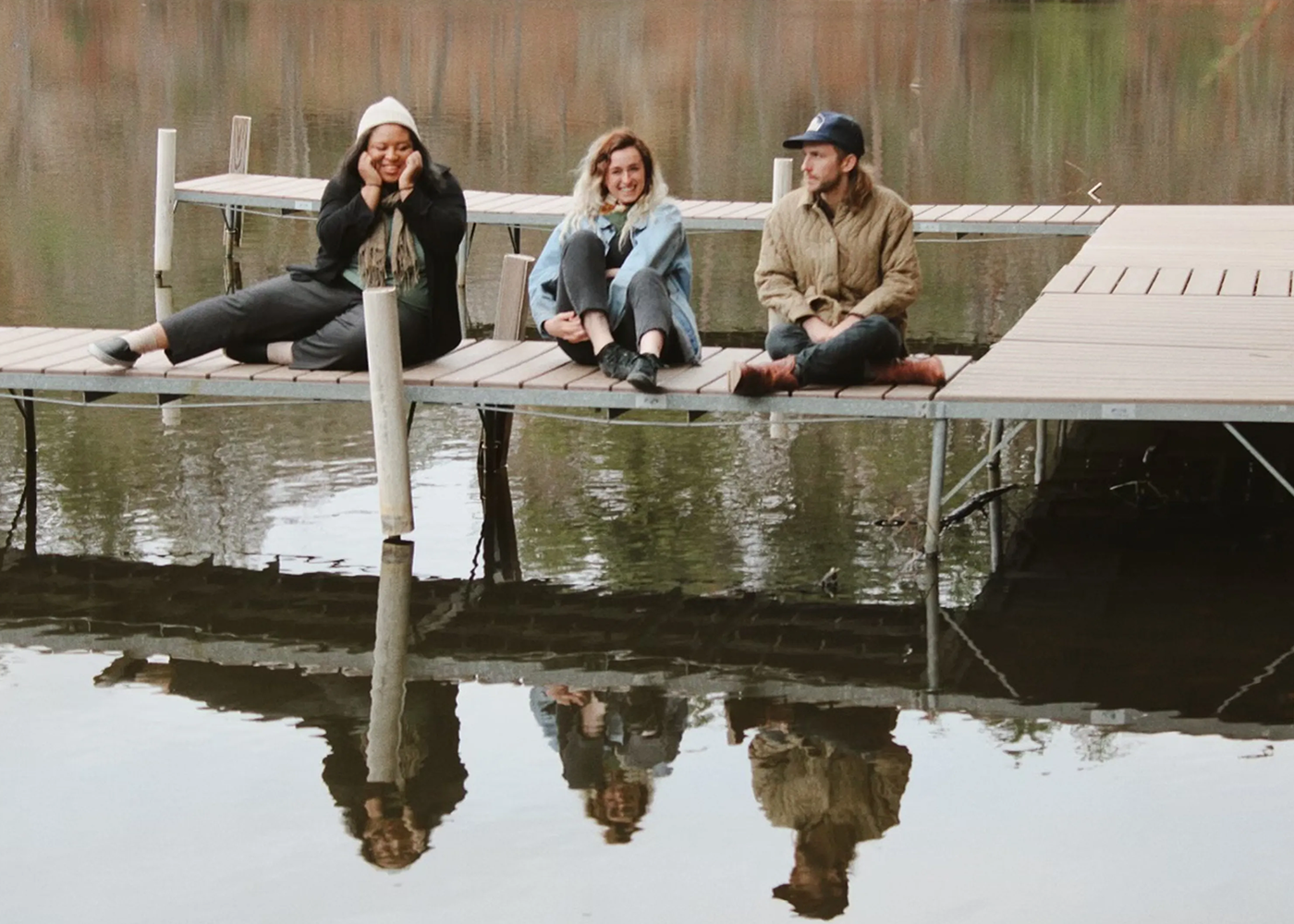 Three people sitting on a wooden dock by calm water with reflections