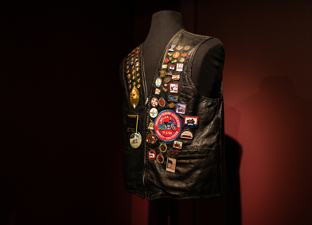 Black leather vest displayed on a mannequin, heavily decorated with pins, badges, and embroidered patches