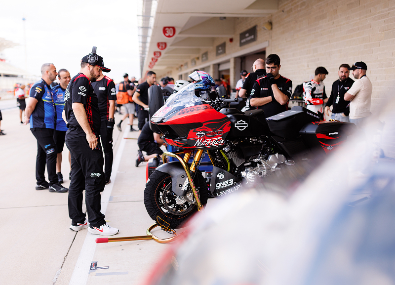 Rider sits on a race motorcycle in the pit lane surrounded by crew and spectators