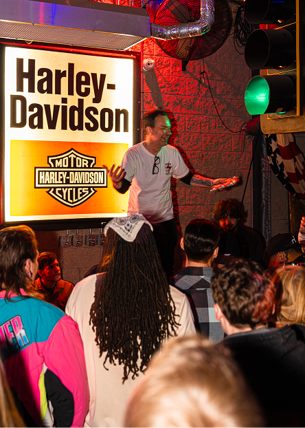 Presenter stands onstage beside a Harley‑Davidson sign addressing a crowd at an indoor event