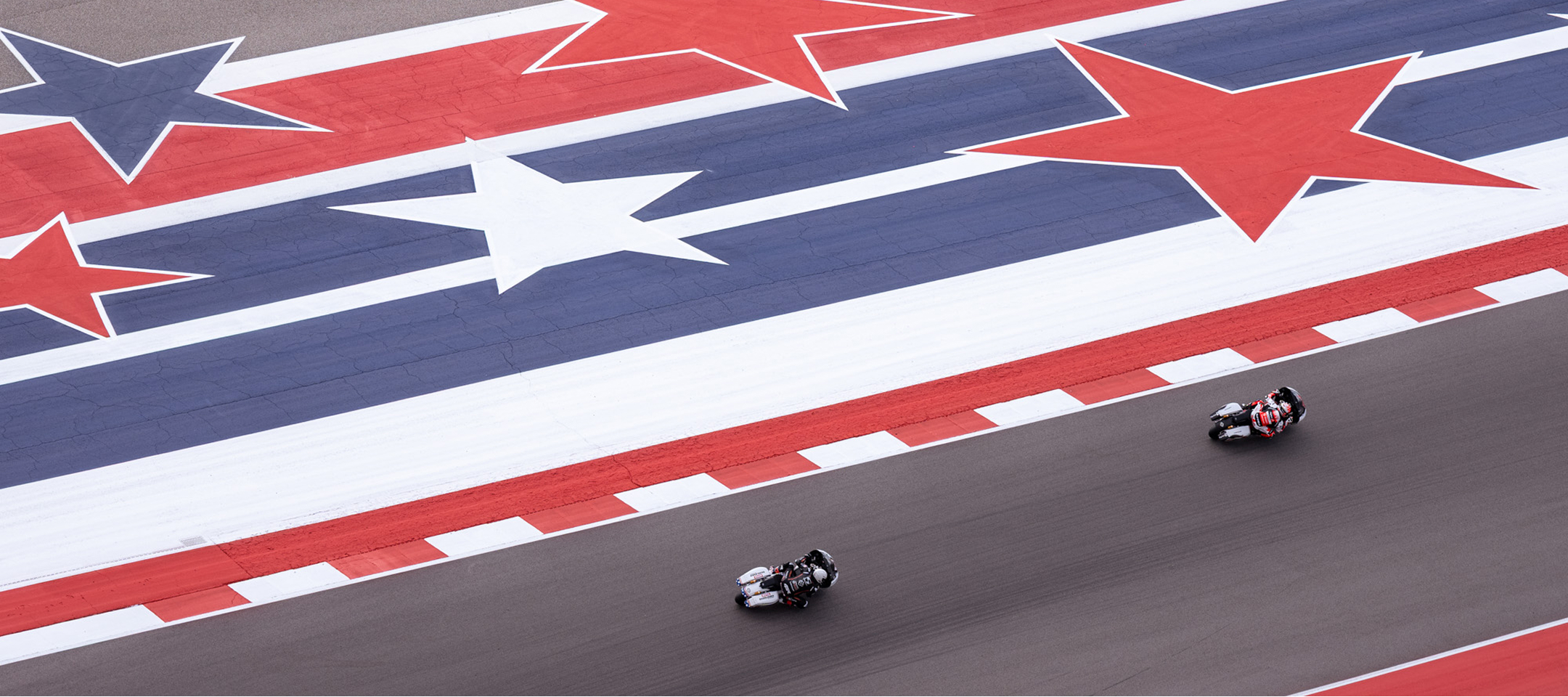 Two motorcycle racers speed down a straightaway beside a large red white and blue star motif painted on the racetrack
