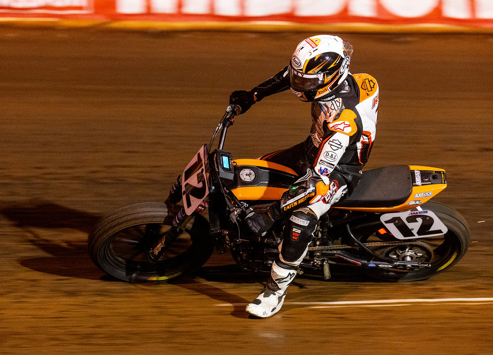 Motorcycle No. 12 charges down the straightaway under lights during a flat track race at night