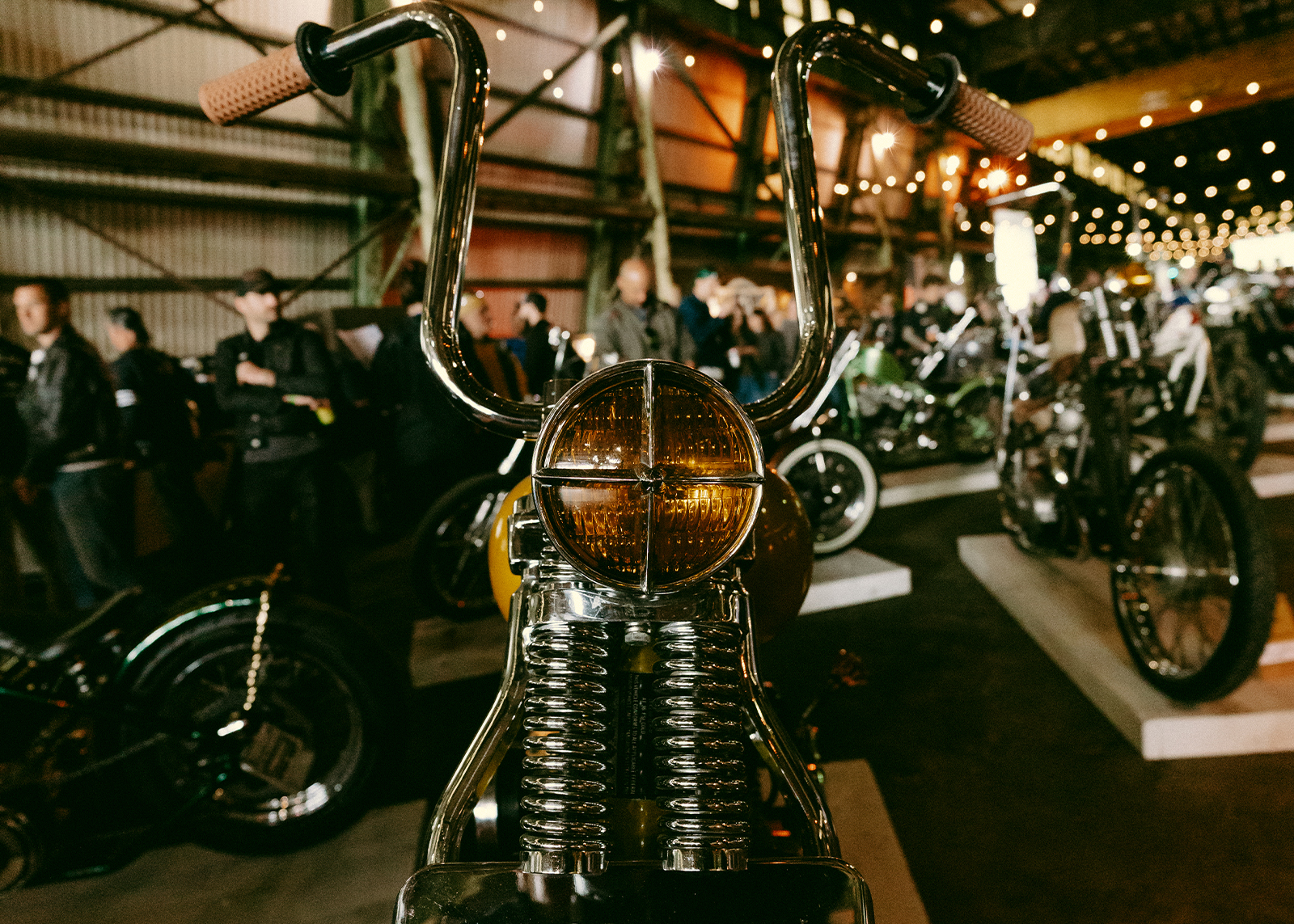 A closeup of a bike, looking straight on into the amber headlight, with a crowd of onlookers in the background.