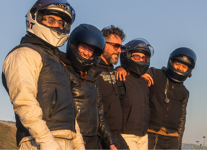 Group of helmeted motorcycle riders standing close together outdoors in warm light with open sky background