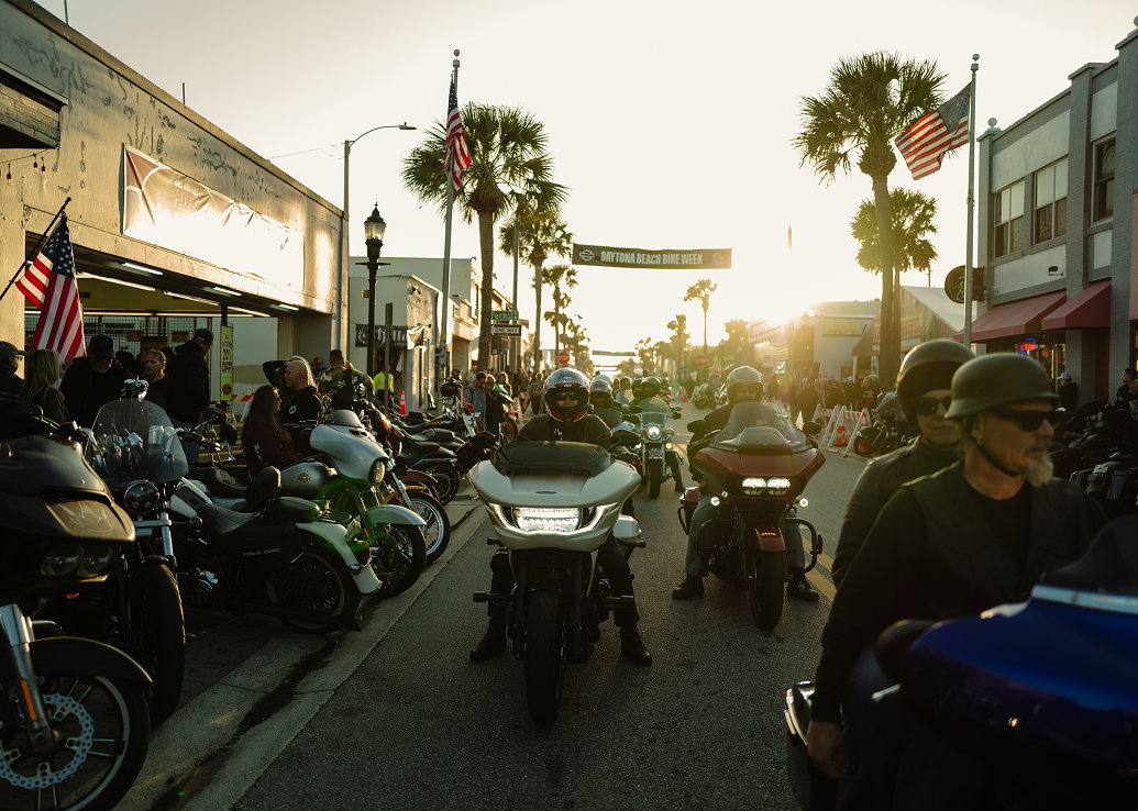 Motorcycles line a busy Daytona Beach main street at sunset, with riders gathered between storefronts and palm trees