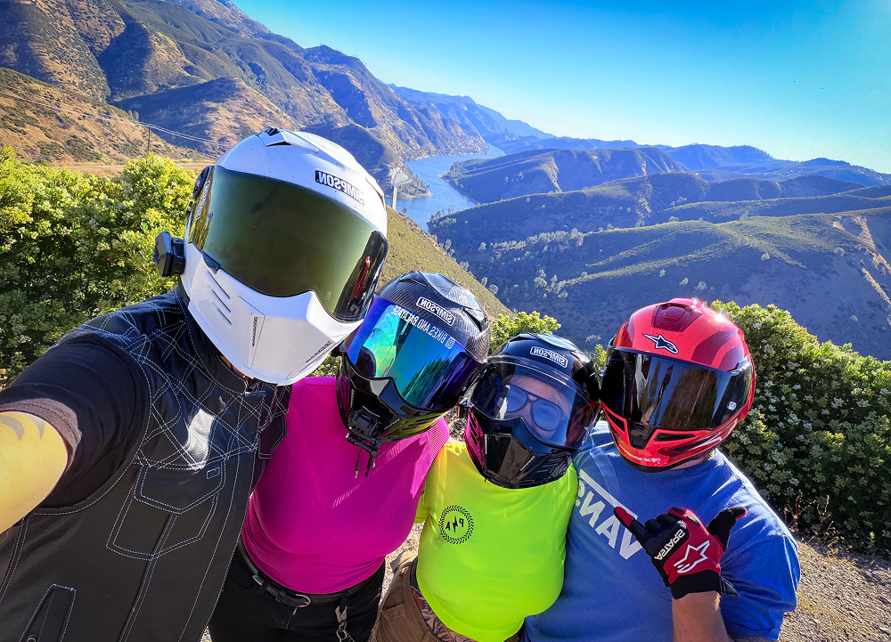 Four motorcycle riders wearing helmets gather for a roadside selfie overlooking winding mountain roads and a reservoir