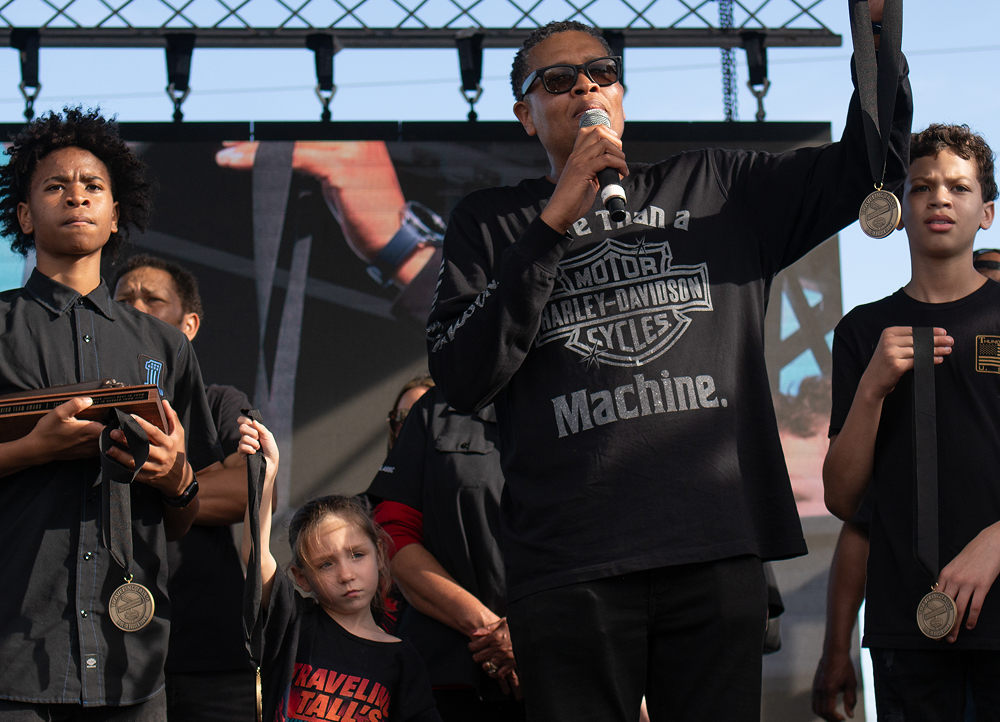 Influencer Traveling Tall speaks into a microphone on stage with his two sons standing beside him