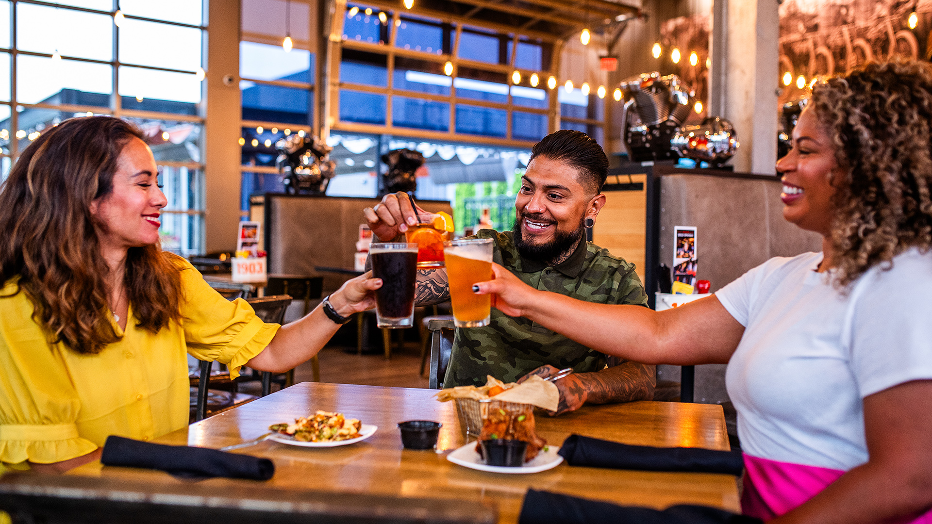 Three people clinking beer glasses at restaurant table with food