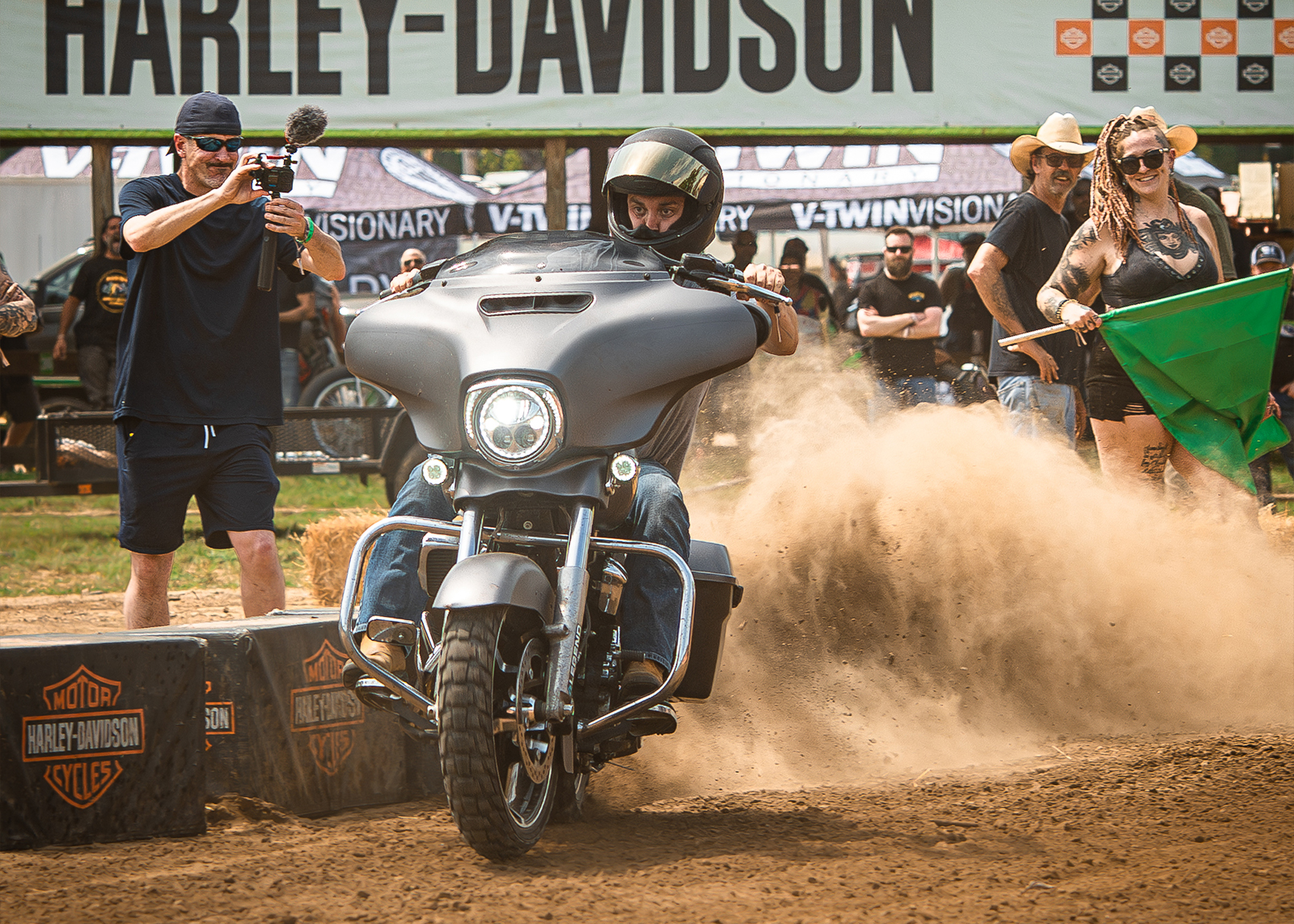 A man on a silver Harley kicks up dirt towards the crowd on the track as he accelerates.