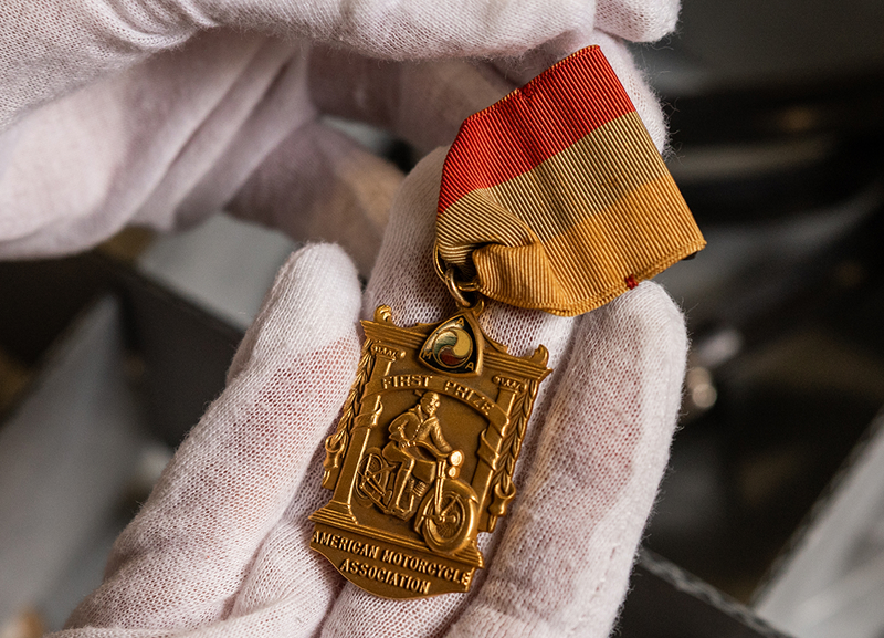 Gloved hands holding a gold medal with red ribbon, featuring embossed motorcycle and event details