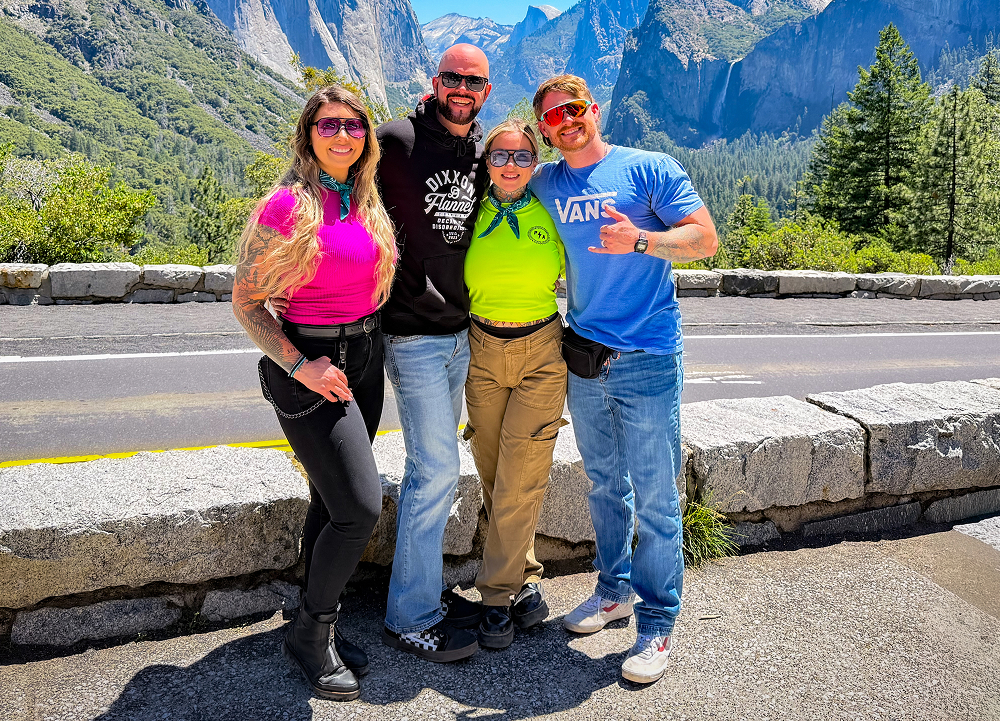 Four travelers pose at a scenic Yosemite Valley overlook, framed by granite cliffs and forested peaks