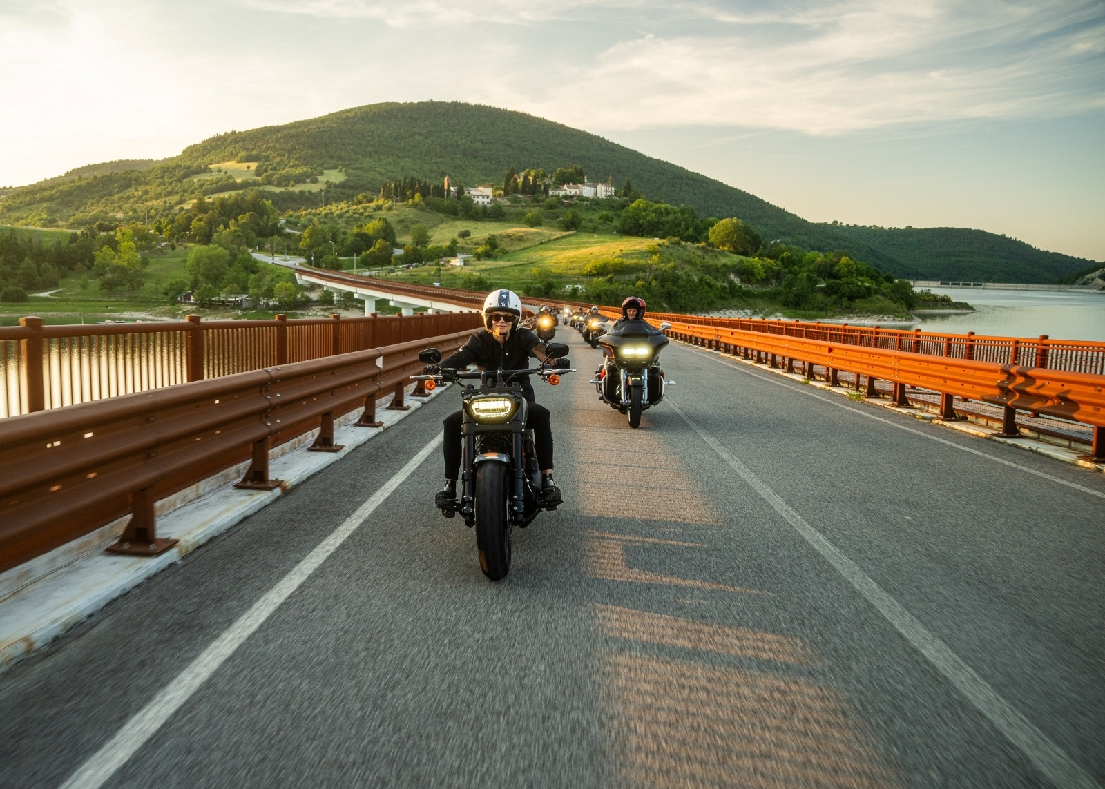 A group of motorcyclists riding across a bridge with hills and greenery in the background.