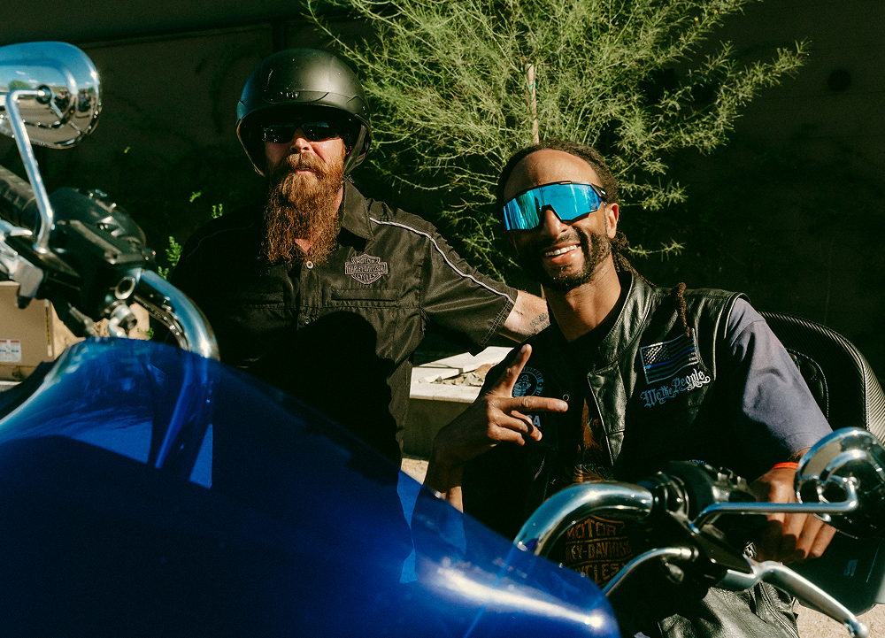 Two motorcycle riders posing beside a blue bike, wearing helmets and sunglasses, parked in shade near trees.
