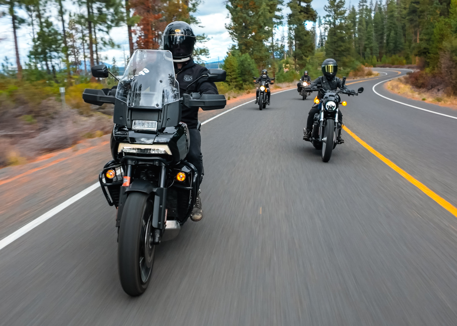 Riders in black helmets cruise together on a winding forest road, surrounded by trees and motion blur.