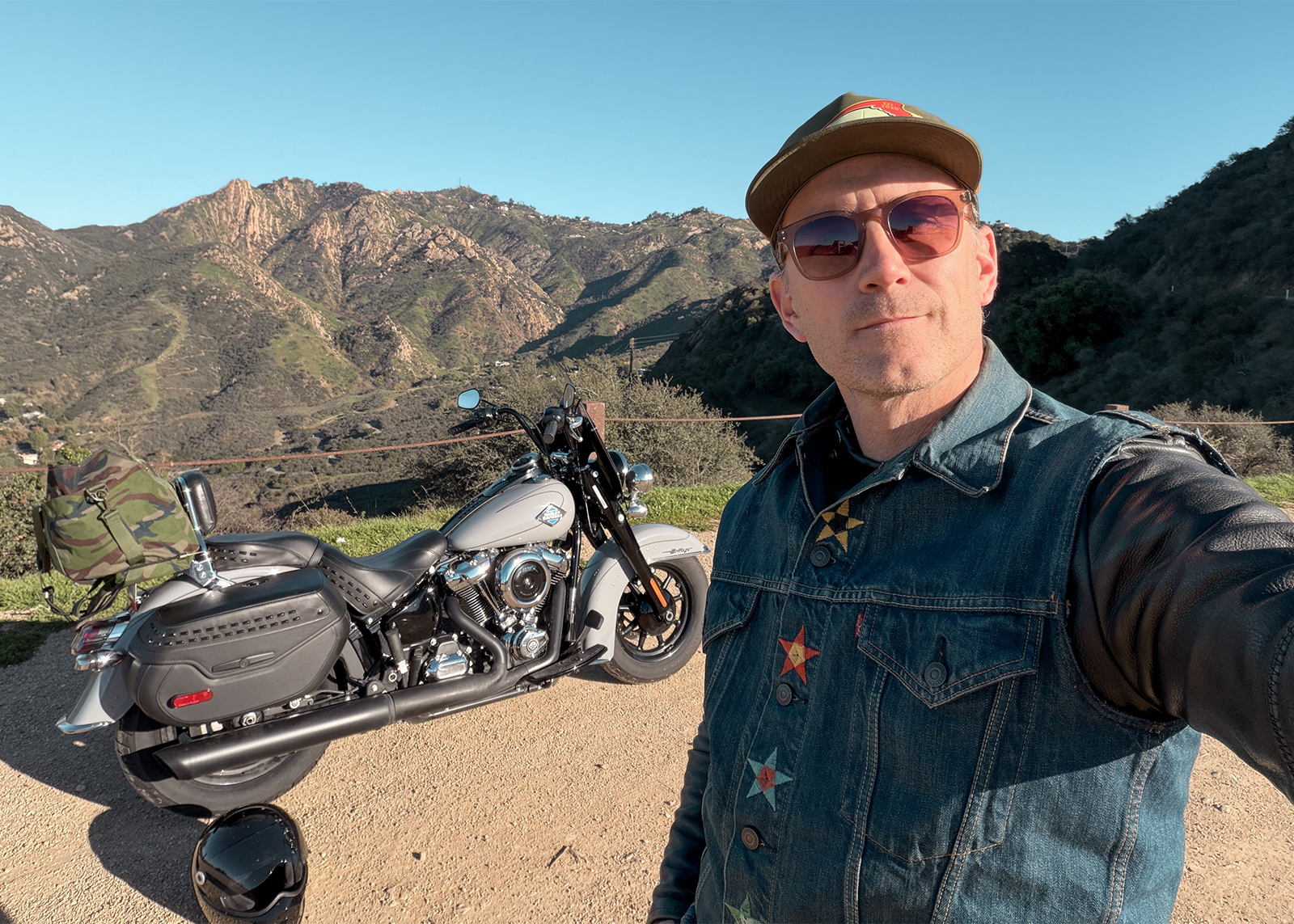 A selfie of the author wearing sunglasses and a denim jacket with his motorcycle and mountains in the background