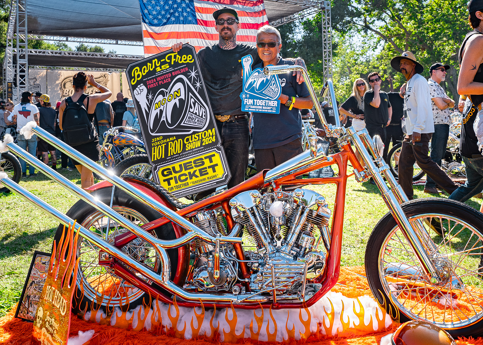 Two men holding up Born Free guest ticket posters behind their custom hot rod, covered in orange and chrome, parked over a bed of plastic flames and an orange rug.