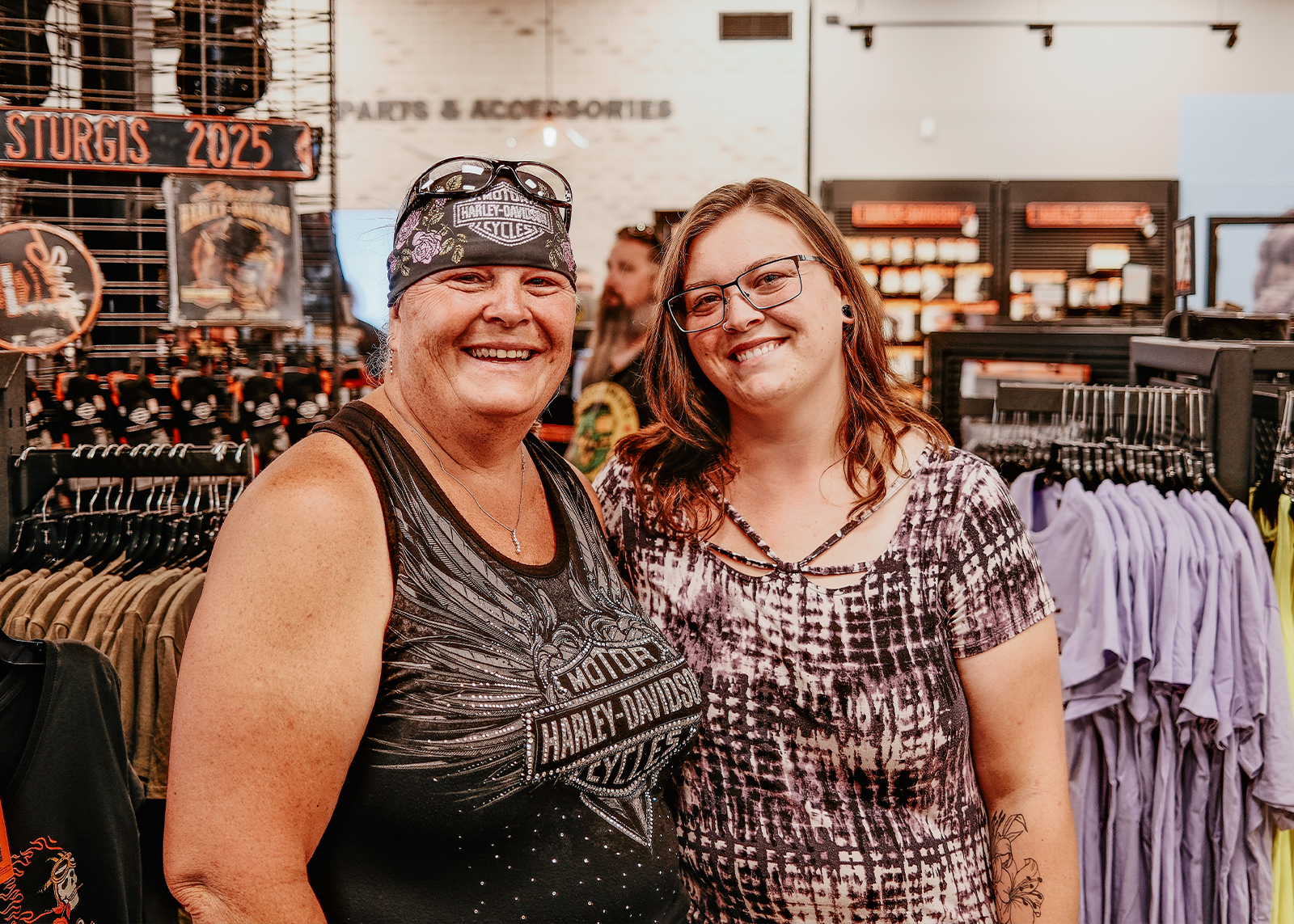 Two smiling women pose inside the Sturgis Harley-Davidson store, surrounded by racks of shirts and accessories.