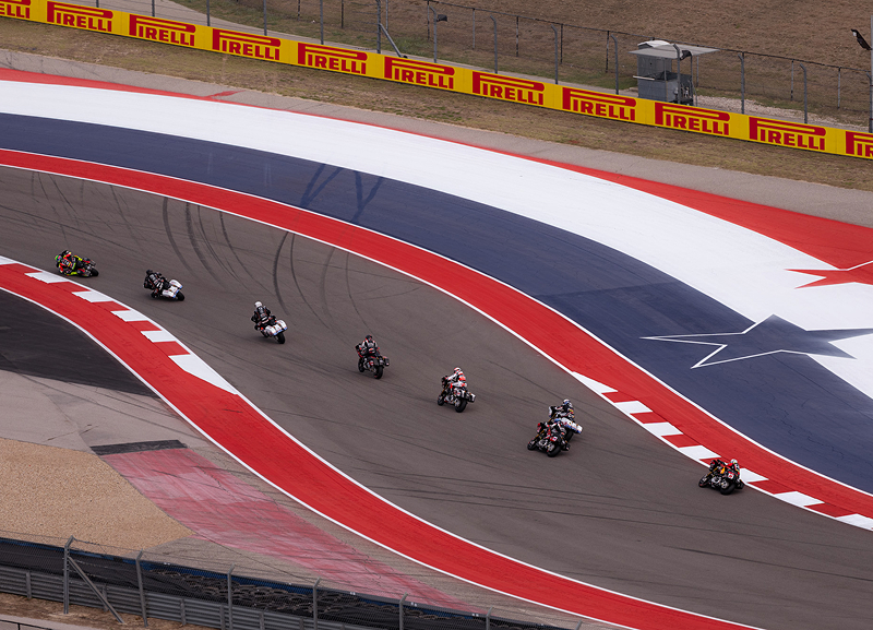 Wide shot of multiple motorcycle racers navigating sweeping red-and-white track curves at speed