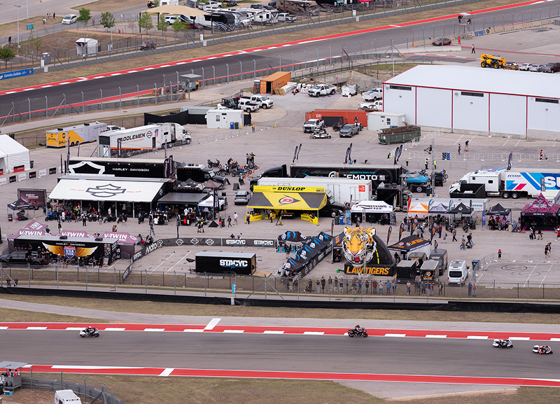 Aerial view of a racetrack paddock with team tents and transporters as motorcycles race past on track below