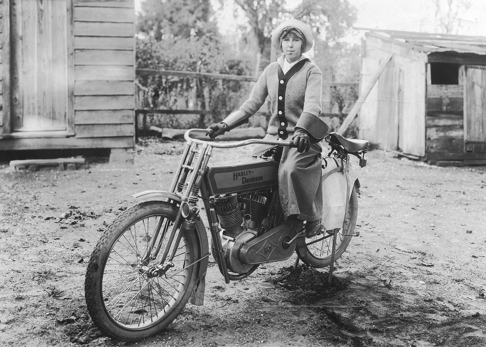 Vintage black-and-white photo of a woman in hat and gloves sitting on a vintage Harley-Davidson motorcycle in a rural yard.