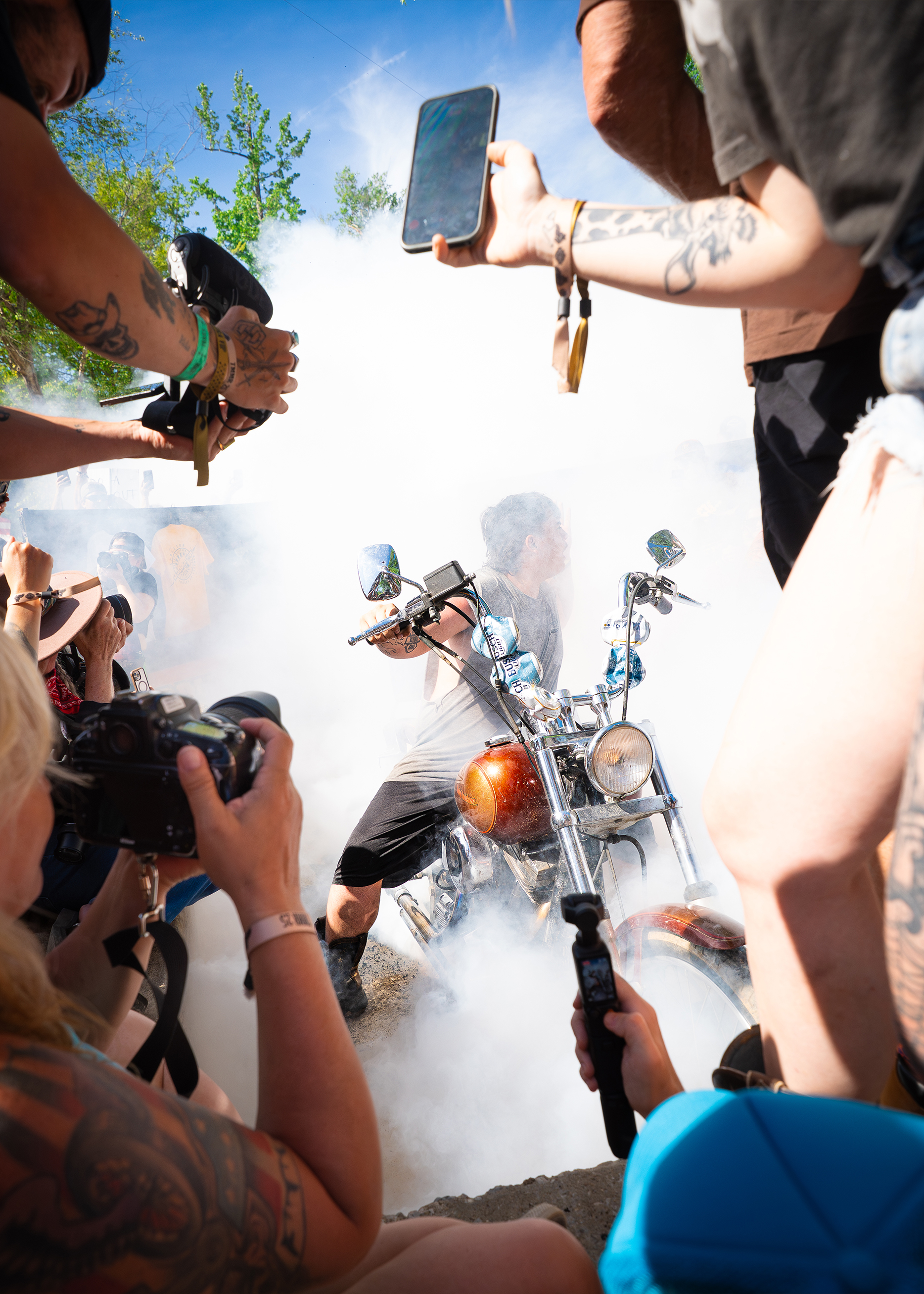A crowd of people lean with their phones and camera to catch the man on his bike in the center hidden by smoke.