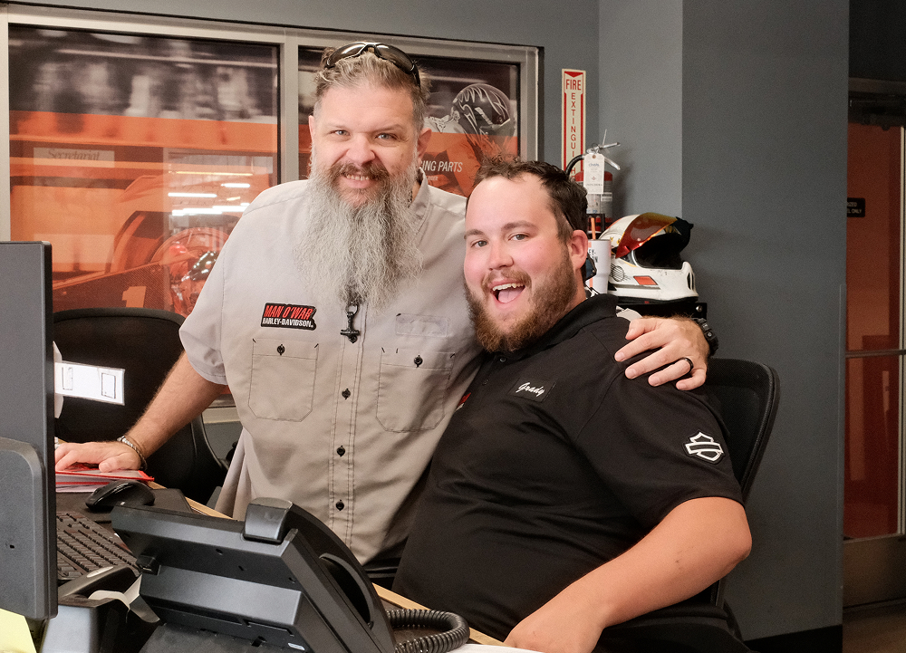 Two smiling dealership employees pose together behind a service counter inside a Harley-Davidson showroom