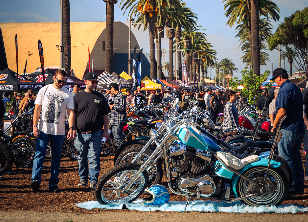 Attendees view custom motorcycles at an outdoor show lined with palm trees, tents, and displays