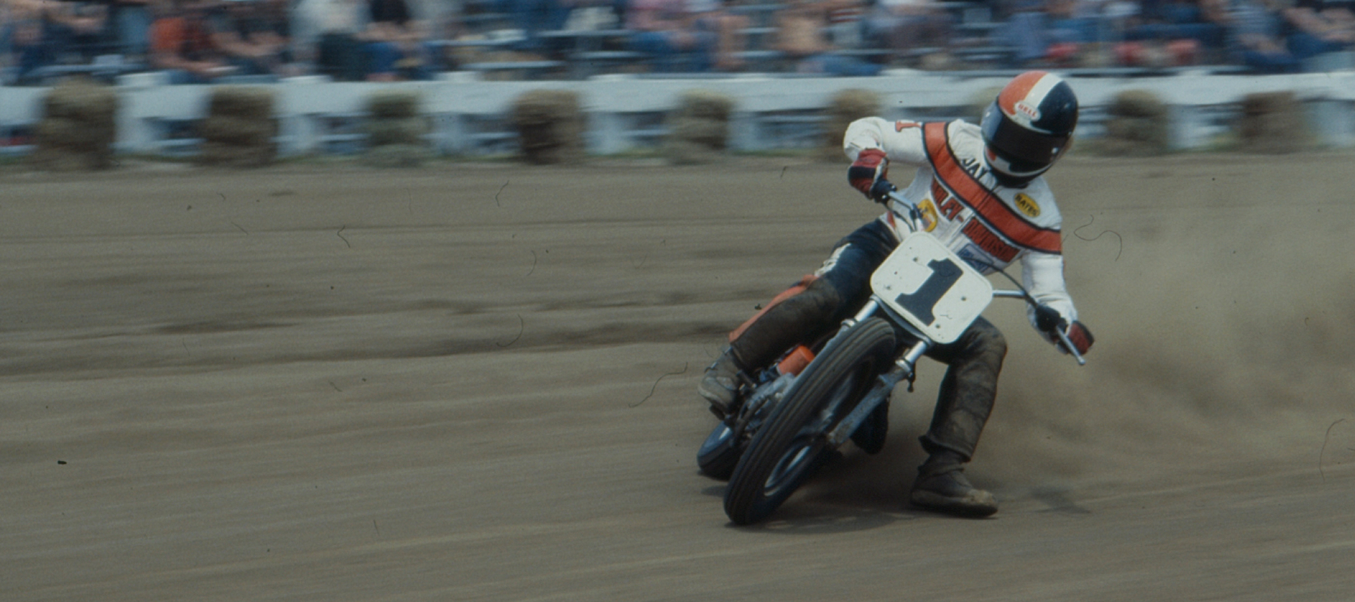 Number 1 rider Jay Springsteen slides a Harley-Davidson flat track motorcycle through a dirt corner as dust flies before spectators