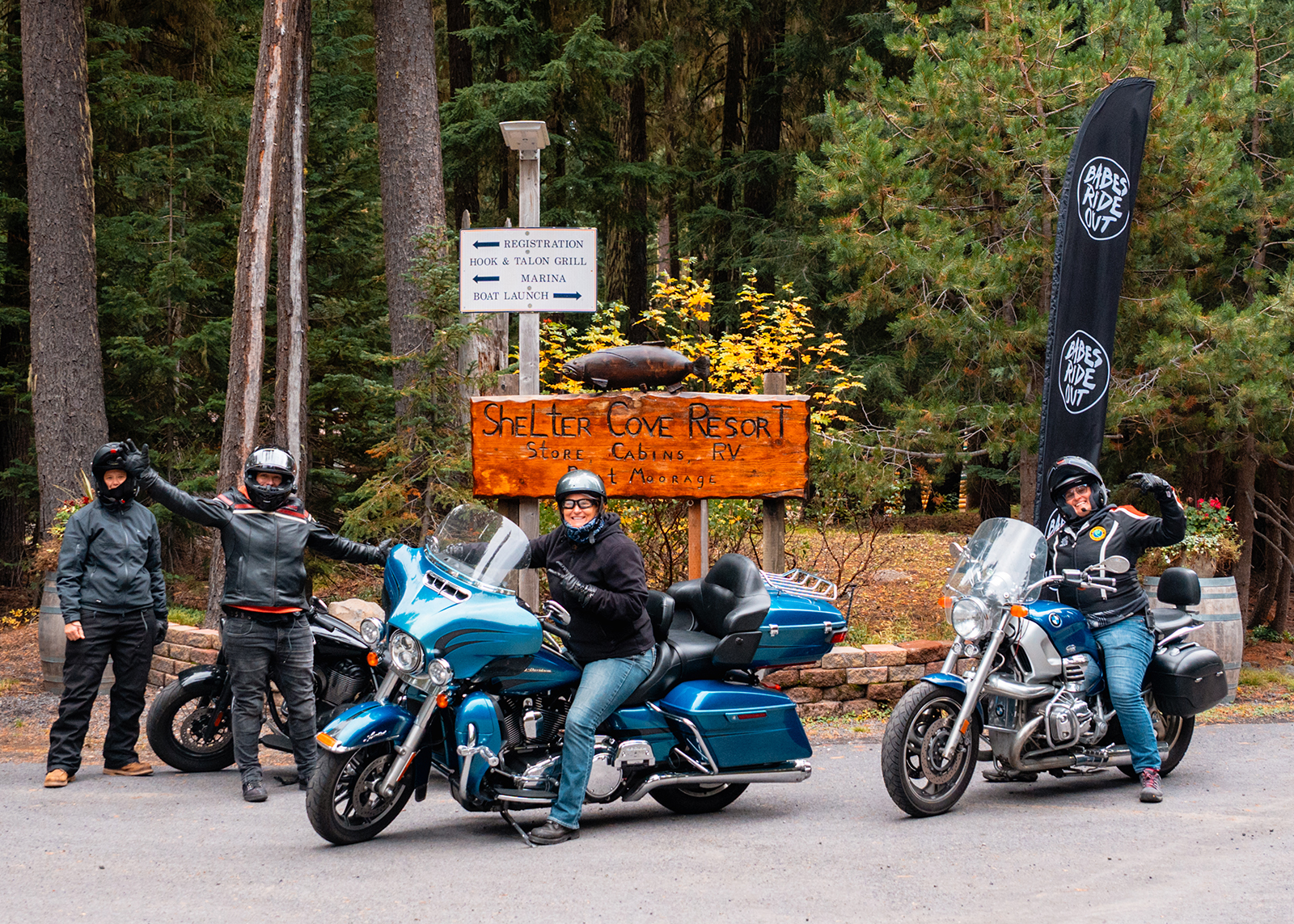 Motorcyclists pose at the Shelter Cove Resort entrance, surrounded by forest and Babes Ride Out banners.