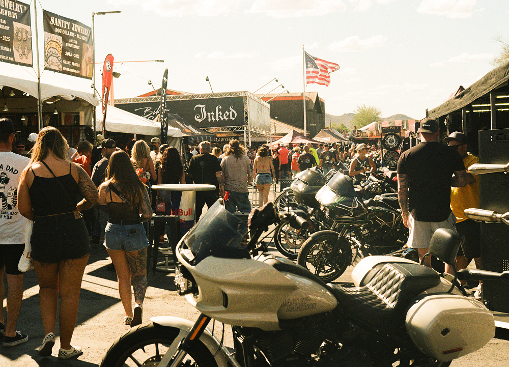 Crowded motorcycle event street filled with parked bikes, vendor booths, flags, and people walking through.