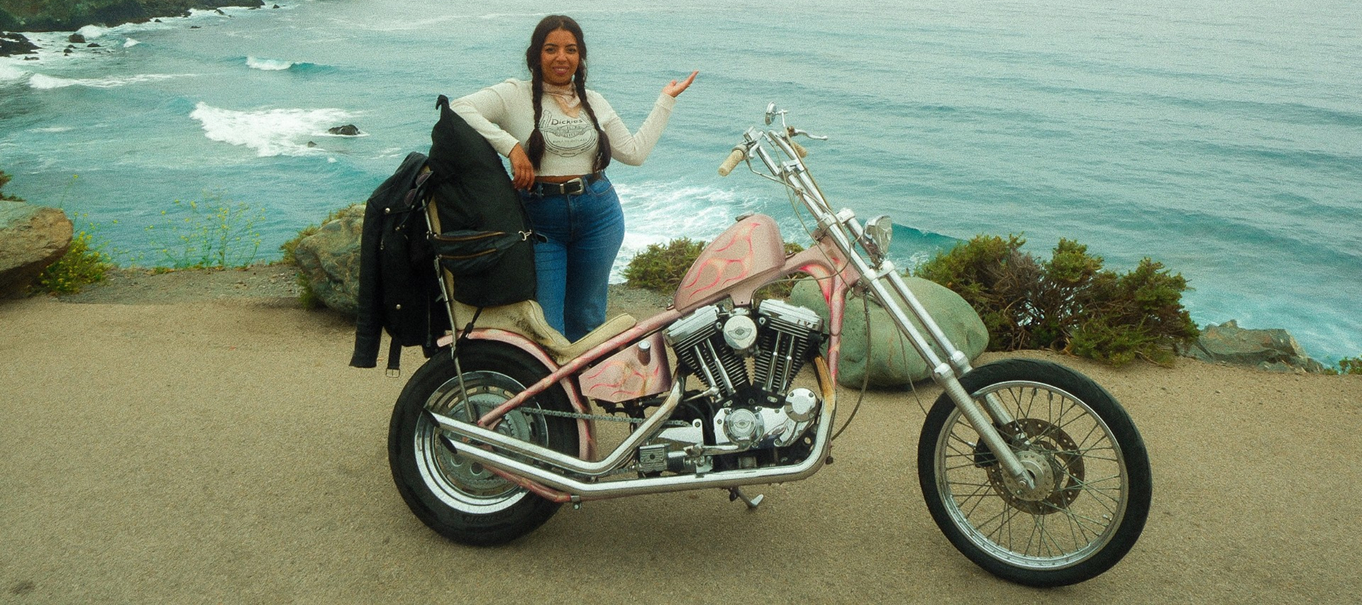 Custom chopper-style motorcycle parked at a coastal overlook with ocean waves, rocky shoreline, and rider beside bike
