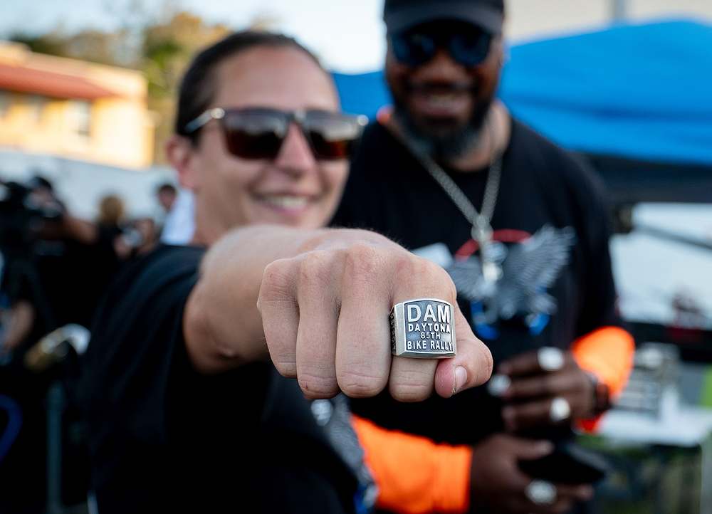 Close-up of a fist wearing a ring that says DAM Daytona 85th Bike Rally extended toward the camera at an outdoor stage event
