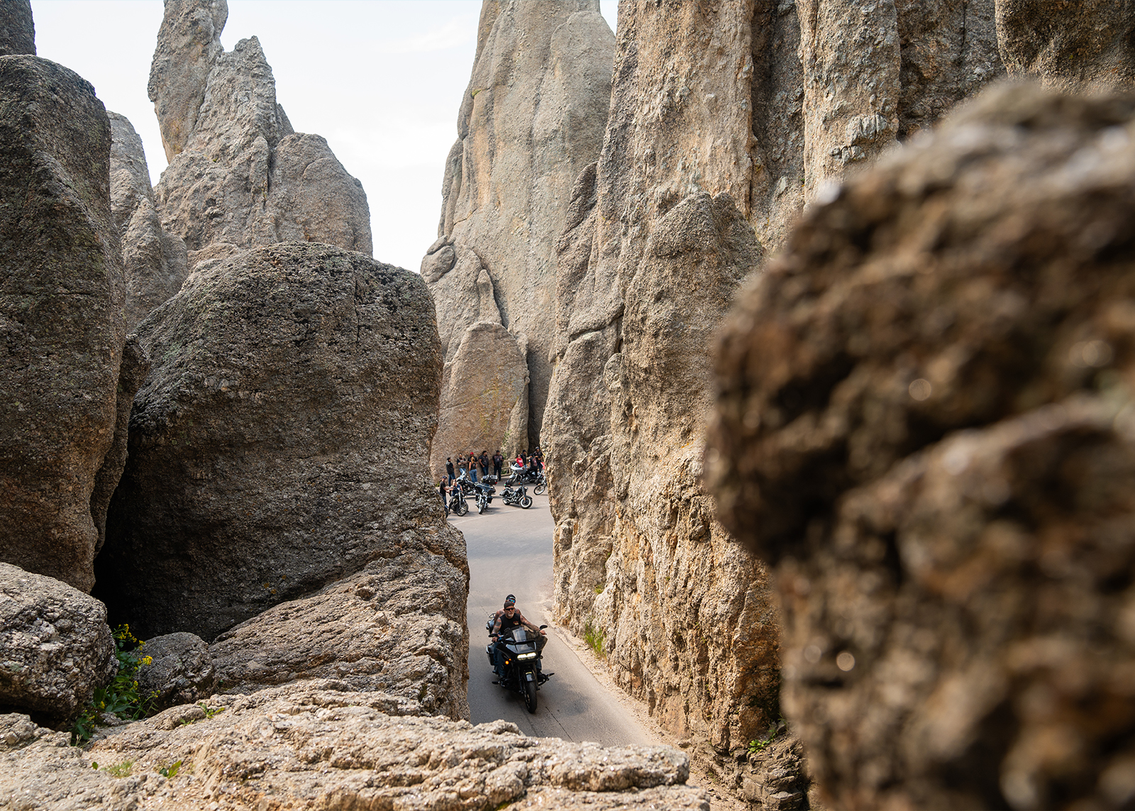 Motorcyclists ride through a narrow mountain pass flanked by towering rock formations in a rugged landscape.