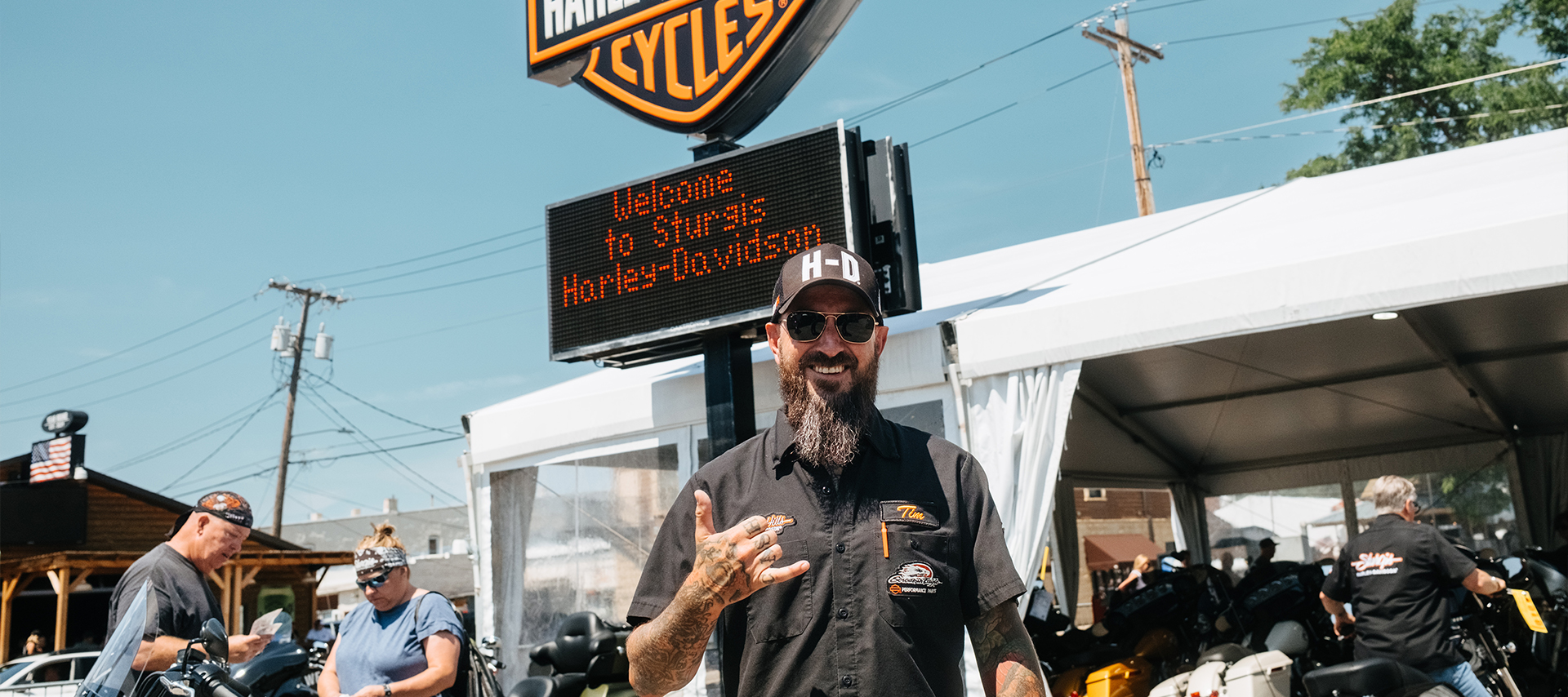 A Sturgis Harley-Davidson dealership staff member with sunglasses, tattoos, and beard smiling at the entrance, flashing a hand sign.