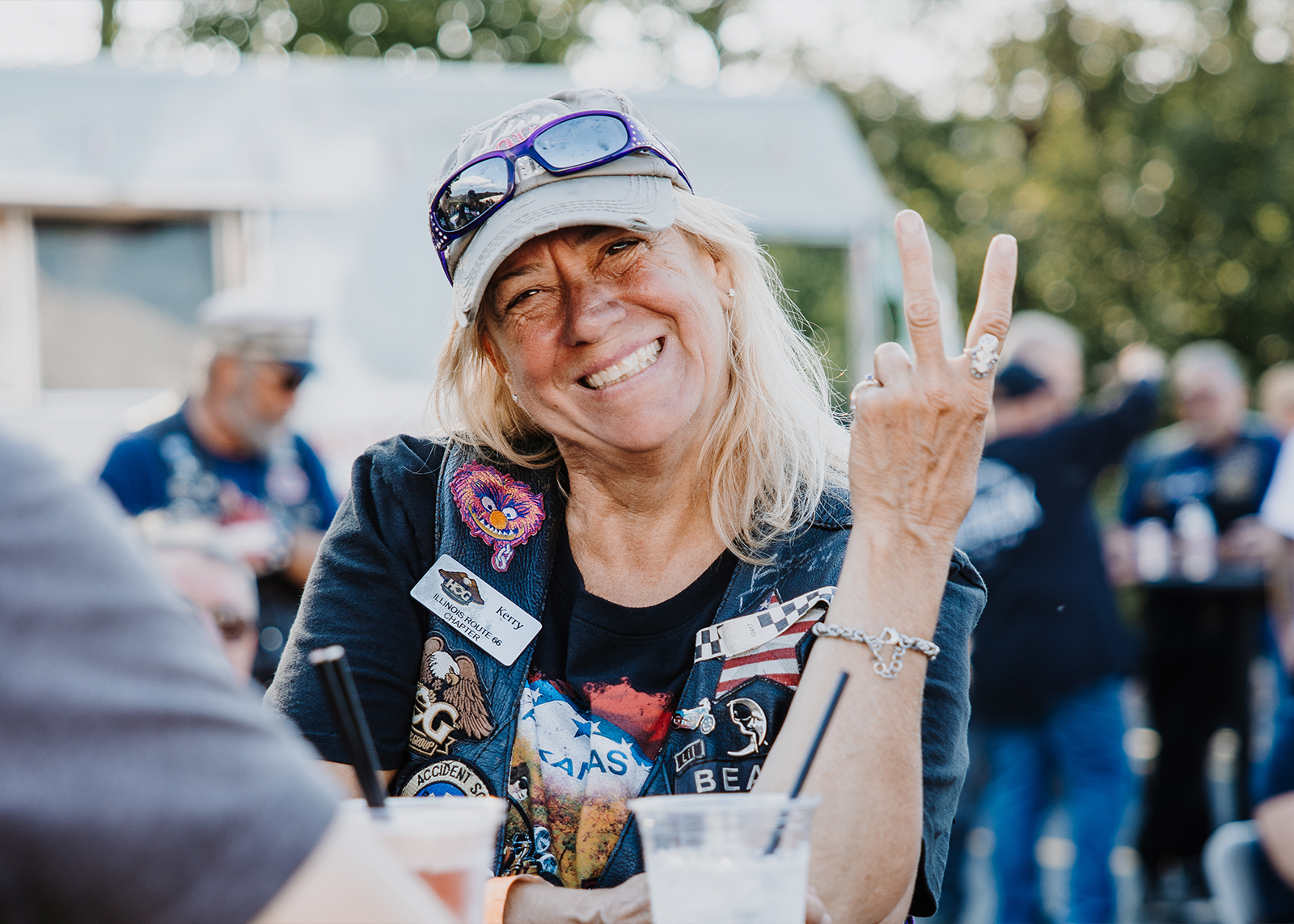 A smiling woman wearing a motorcycle vest covered in patches flashes a peace sign to the camera