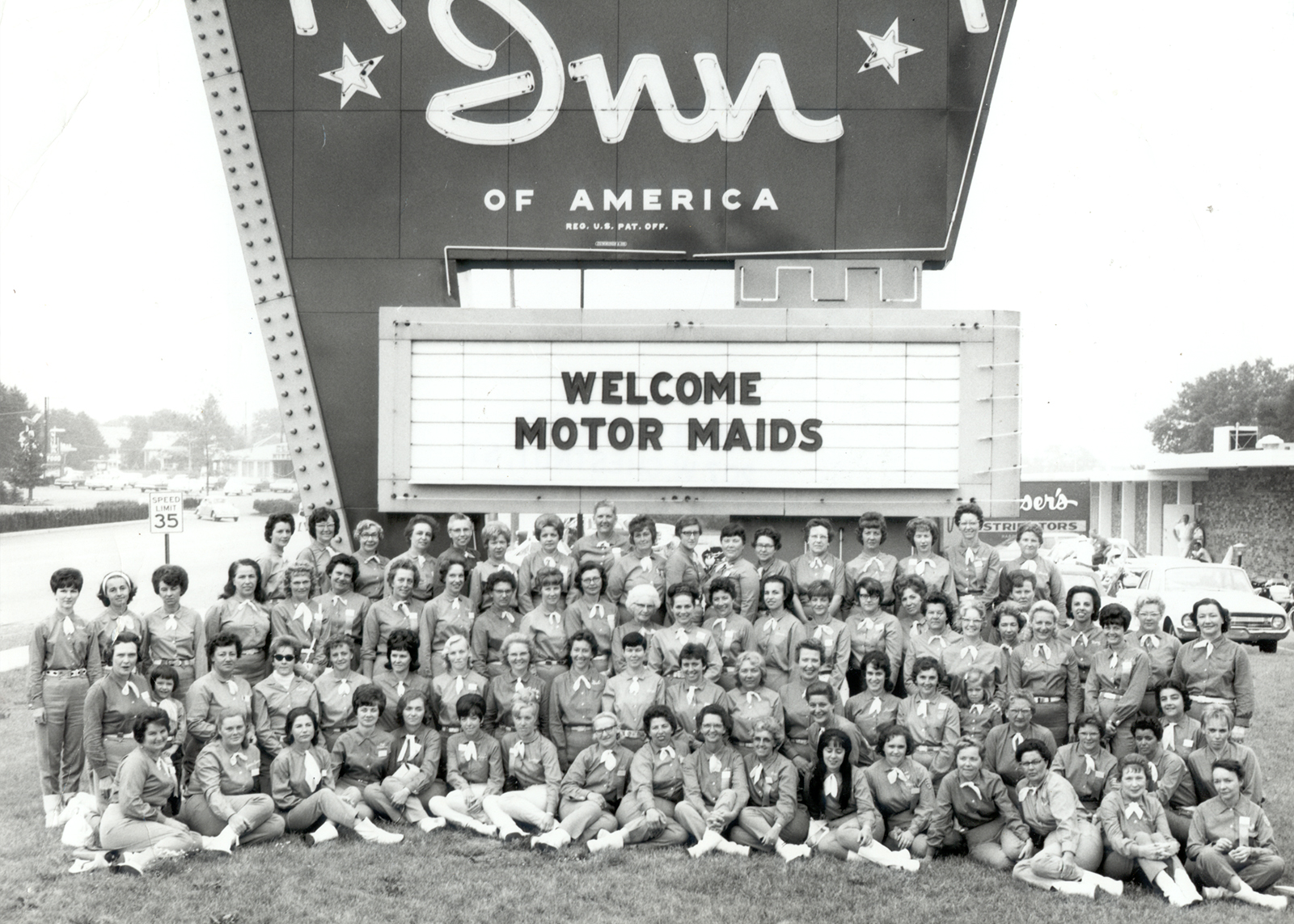 A vintage black and white group photo of the Motor Maids in front of a large Holiday Inn sign and marquee that says “Welcome Motor Maids” during a rally in 1967.