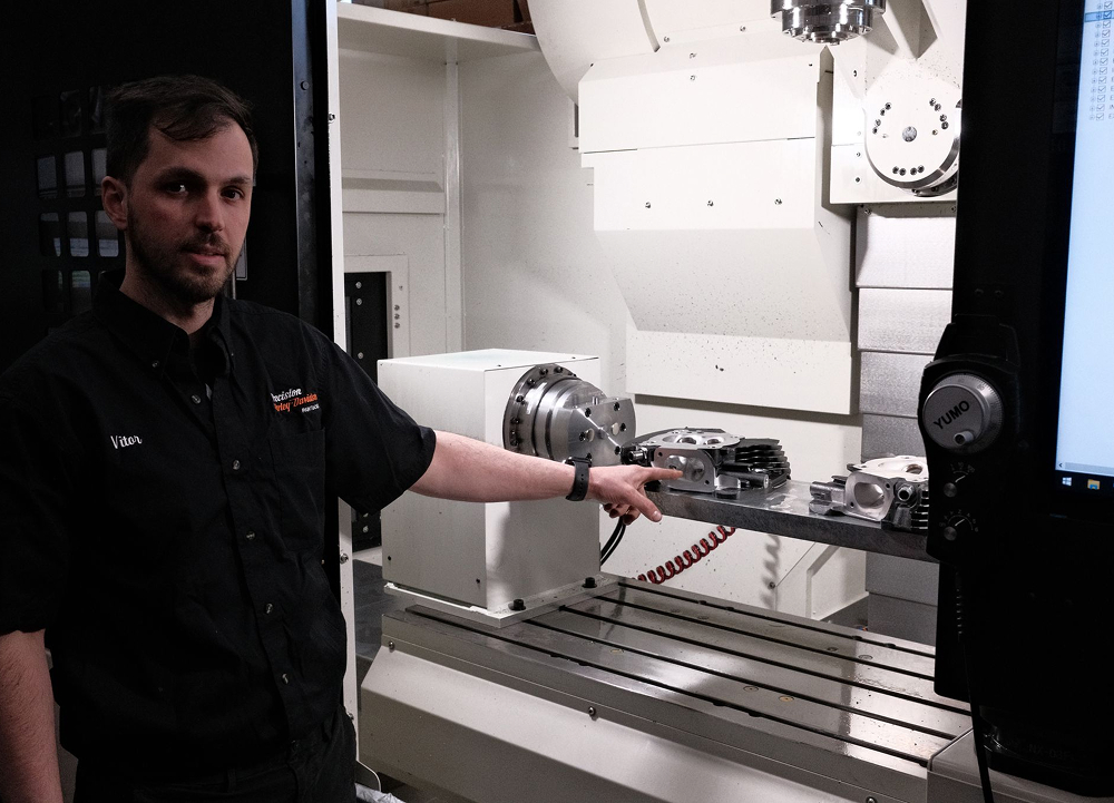 Precision H-D Master Tech Vitor Raposo standing and pointing towards a machined engine part sitting on a cnc machine