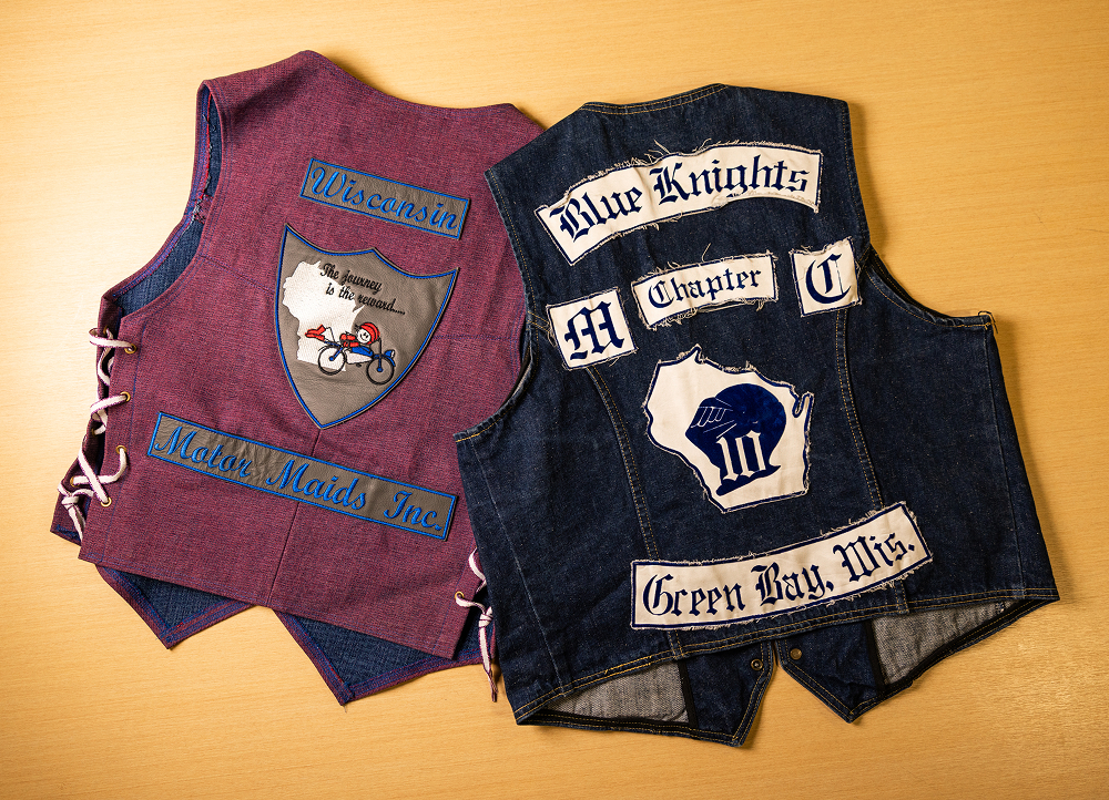 Two sleeveless denim and fabric vests laid on a table, displaying stitched patches, text labels, and club-style emblems
