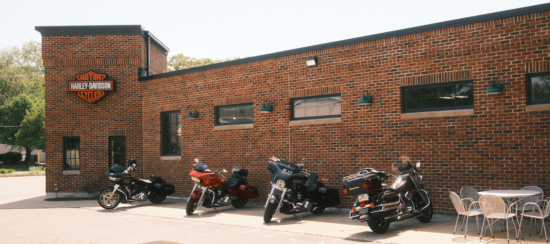 Row of parked Harley-Davidson motorcycles outside brick dealership with logo sign and patio seating