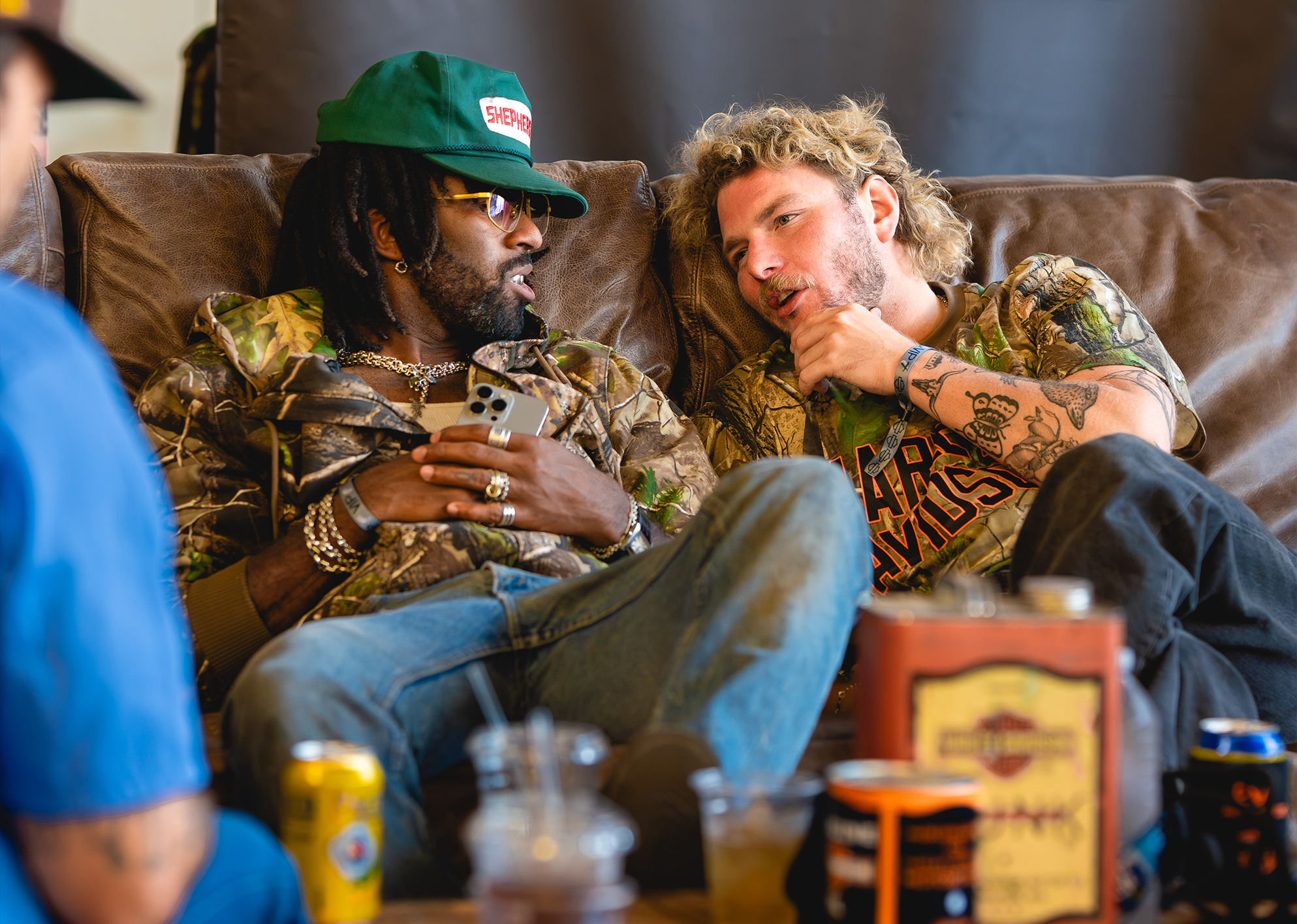 Two men in Harley-Davidson x Realtree camo gear relax on a couch talking amid drinks and friends.