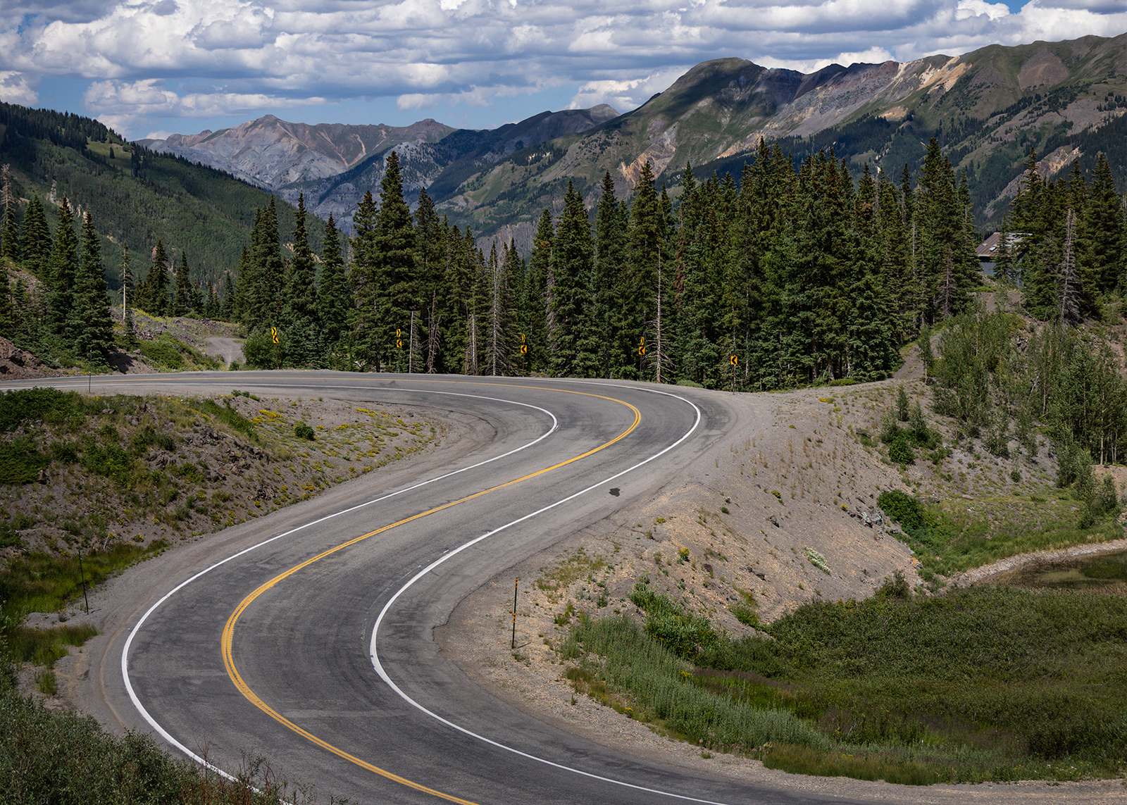 Curving mountain highway winds through pine forest with rugged peaks in the background under clouds.