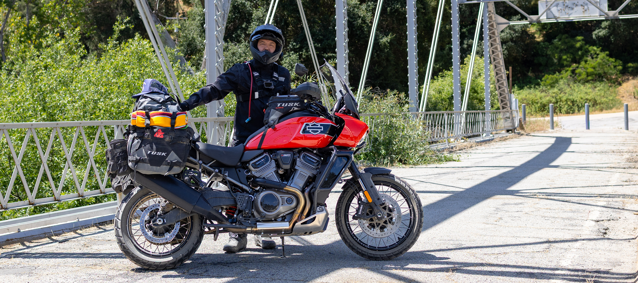 Red H-D Pan America motorcycle parked on a steel bridge with fully loaded luggage, rider standing beside it on sunny day