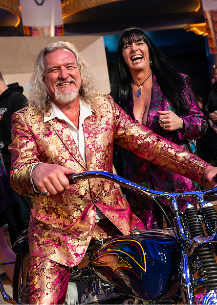 Person in a patterned suit posing on a custom motorcycle inside a crowded event space