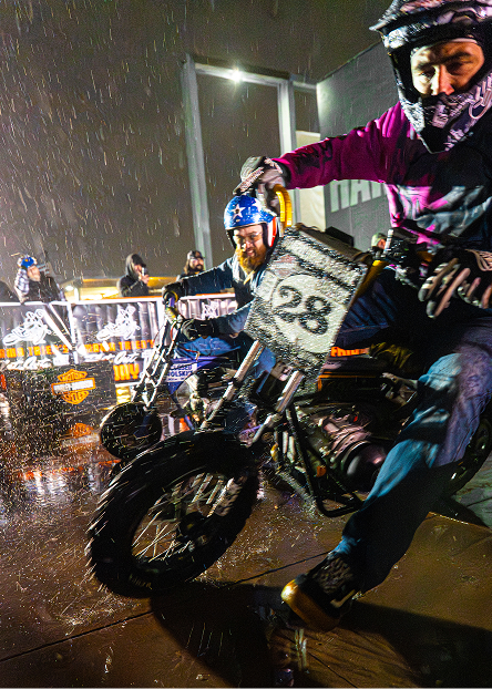 Rider on a small motorcycle navigates a wet track during a rainy nighttime race surrounded by spectators