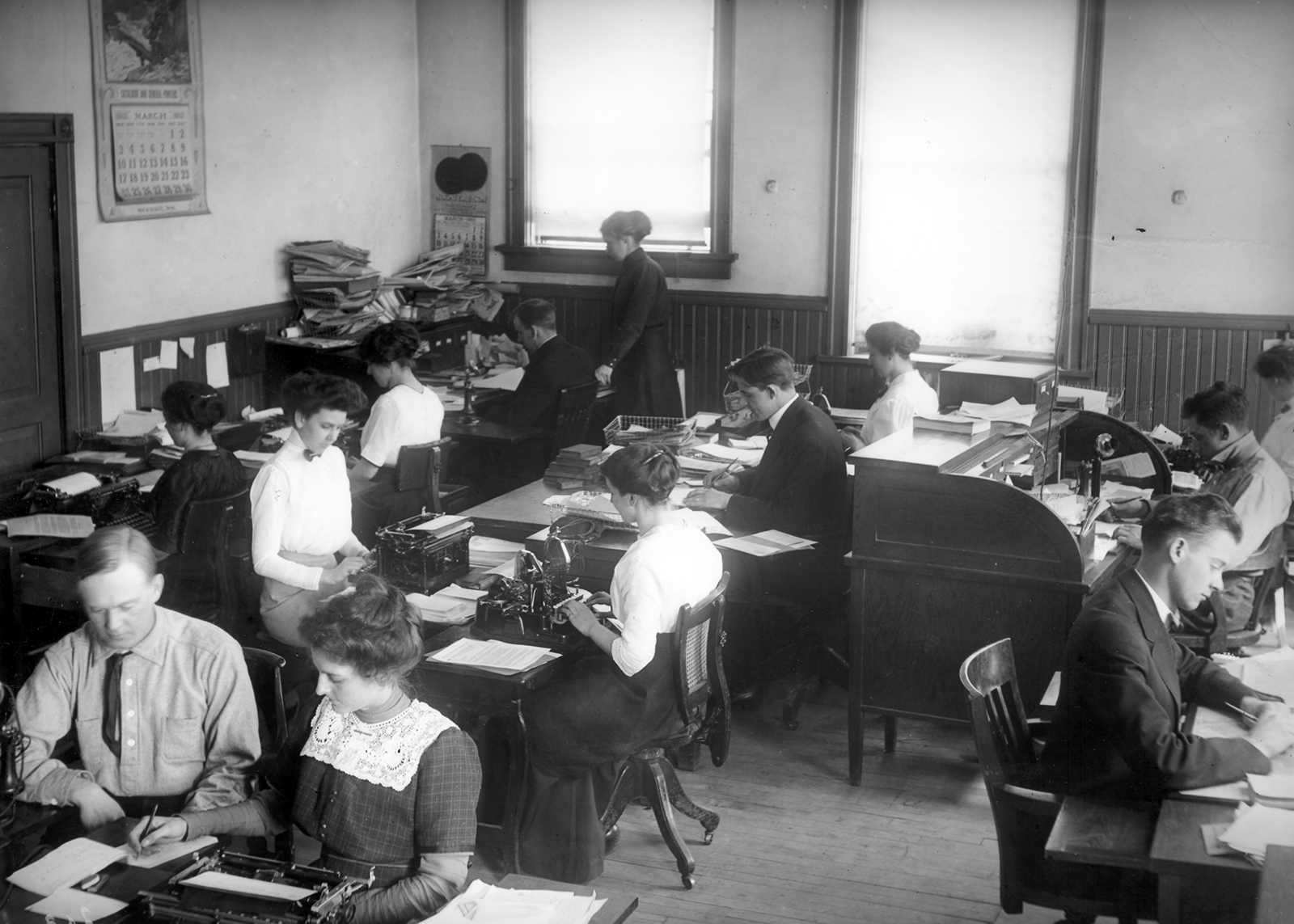 Many office workers sit in a room typing and writing