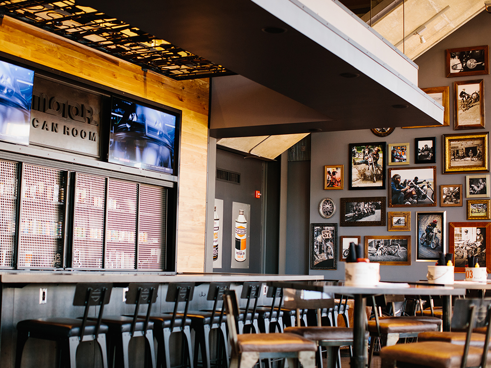 Empty bar seating area with tall stools, wall photo gallery, and large display screens