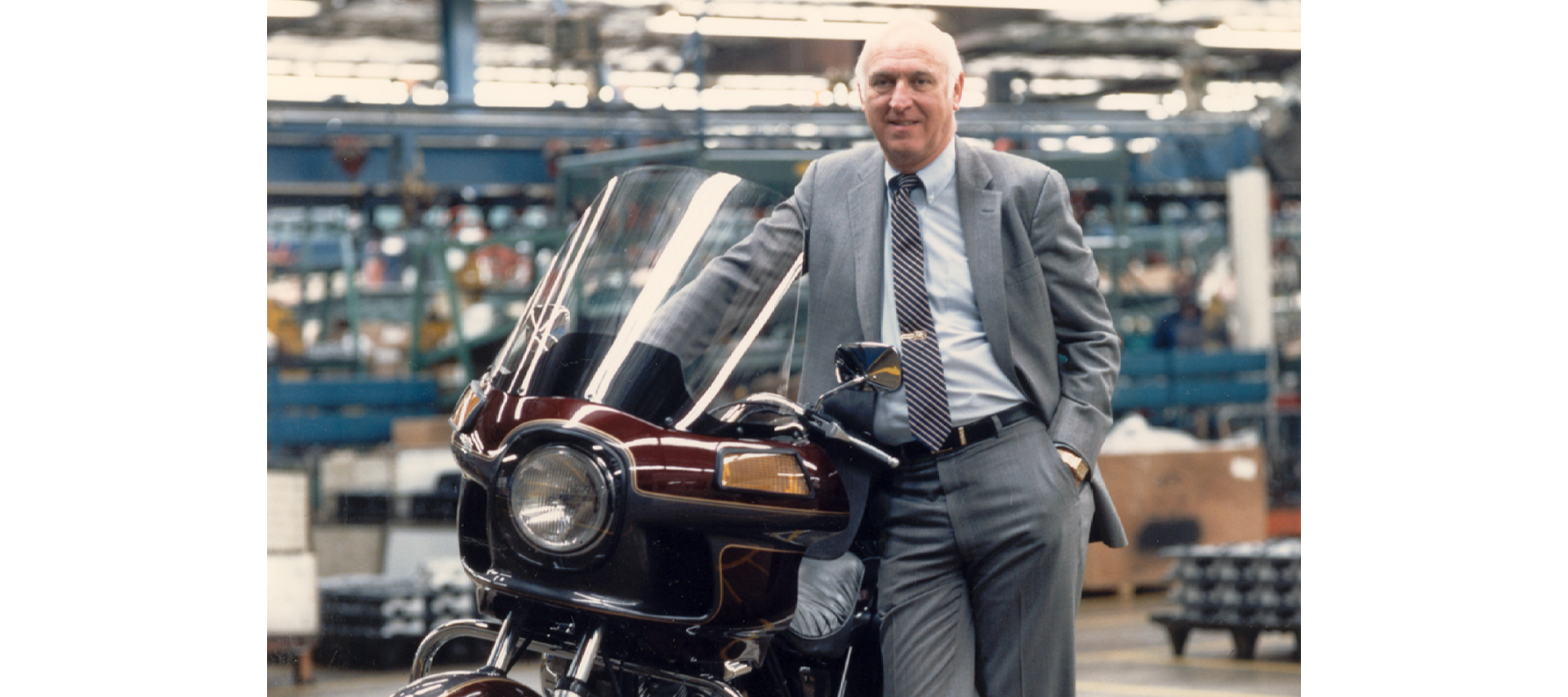 A man in a suit poses with a motorcycle on a factory floor